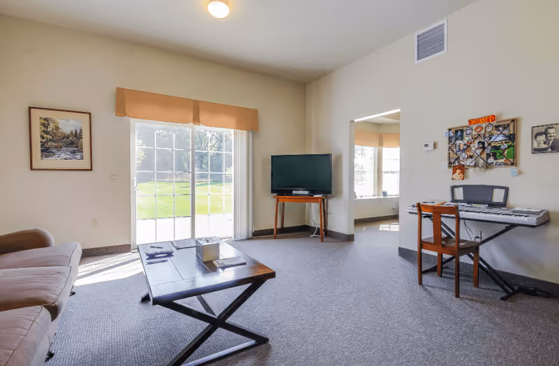 A bright living room with a beige sofa on the left, a wooden coffee table in the center, and a flat-screen TV on a small wooden stand against the far wall. There is a sliding glass door with a beige valance letting in natural light and showing a green outdoor area. On the right side, there is a keyboard on a stand with a wooden chair in front of it, and a bulletin board with photos and notes on the wall above the keyboard. The room has light-colored walls and carpeted flooring.