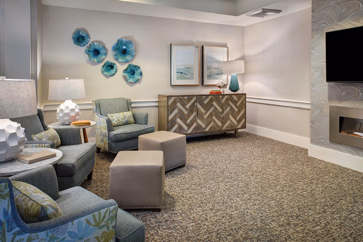 A cozy lounge room with upholstered armchairs, ottomans, side tables, decorative wall art, a chevron-patterned credenza, and a wall-mounted TV with fireplace.