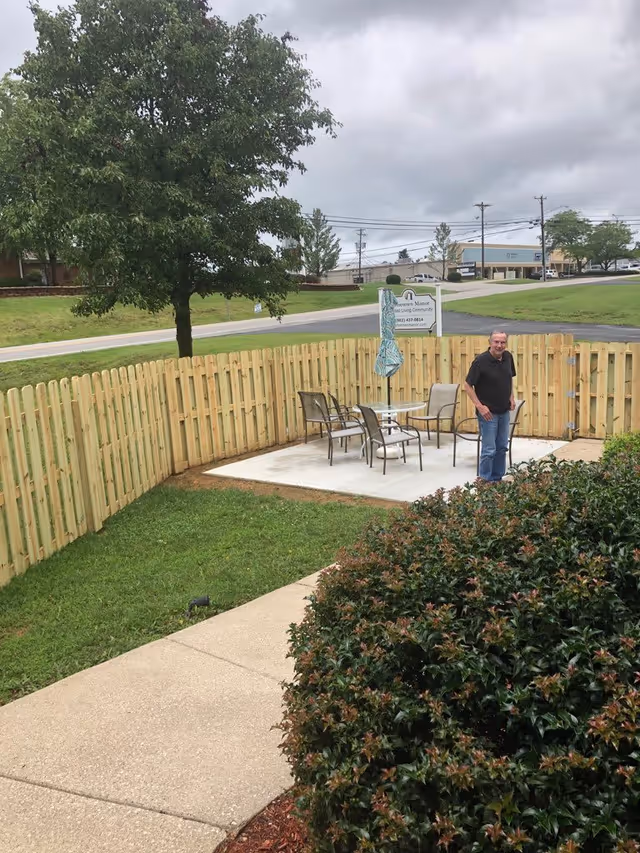An outdoor fenced patio area with a concrete slab, a glass table with four chairs, and a closed umbrella. A man stands near the table. There is a tree and a bush in the foreground, and a road with buildings and power lines in the background under a cloudy sky.