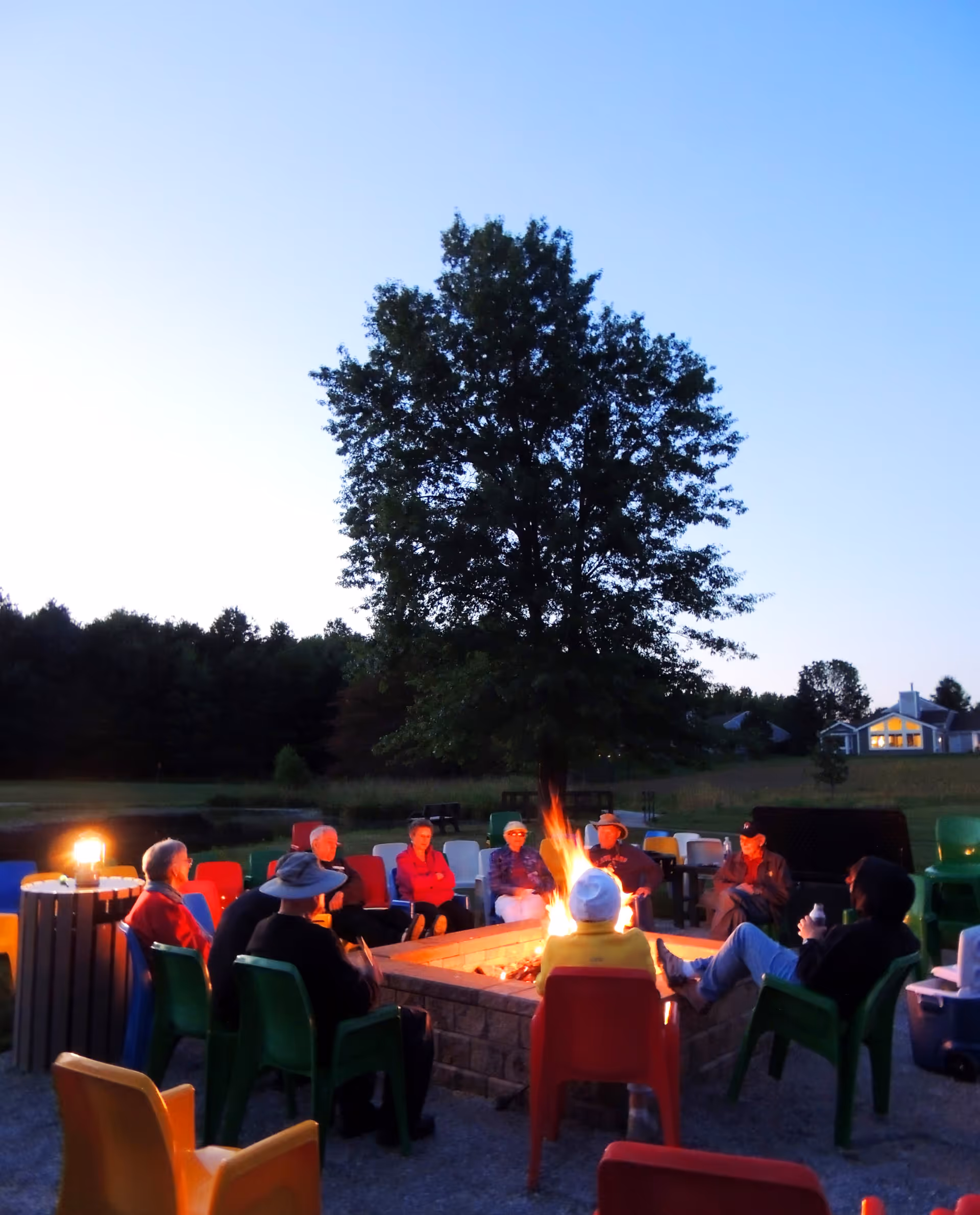 A group of elderly people sitting around a fire pit outdoors in colorful chairs during dusk, with a large tree and a house visible in the background.