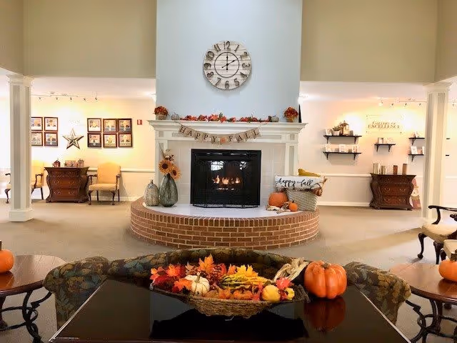 A cozy living room area with a brick fireplace decorated for fall with pumpkins, autumn leaves, and a banner that reads 'Happy Fall'. Above the fireplace is a large round clock. The room has beige walls and carpet, with chairs and wooden furniture arranged around the fireplace. Shelves and framed pictures decorate the walls in the background.