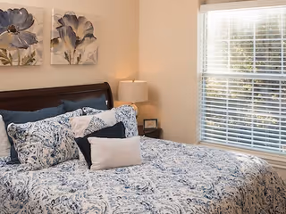 A cozy bedroom with a bed featuring a blue and white patterned comforter and multiple pillows. There is a wooden headboard, a nightstand with a lamp and a small framed picture, and two floral paintings on the wall above the bed. A window with white blinds allows natural light into the room.