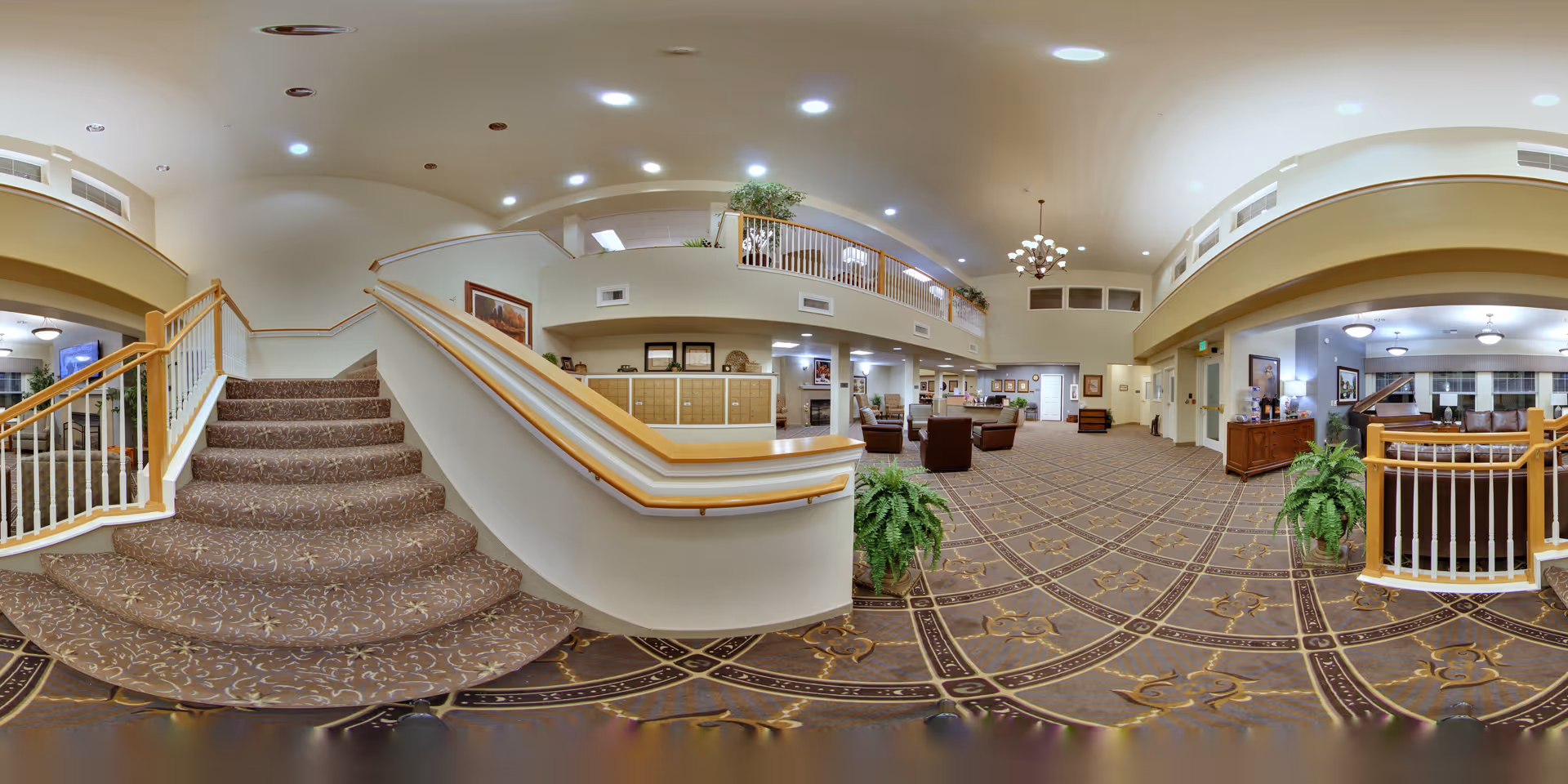 Spacious senior living lobby with a carpeted central staircase, seating areas, balcony overlook, and decorative plants.