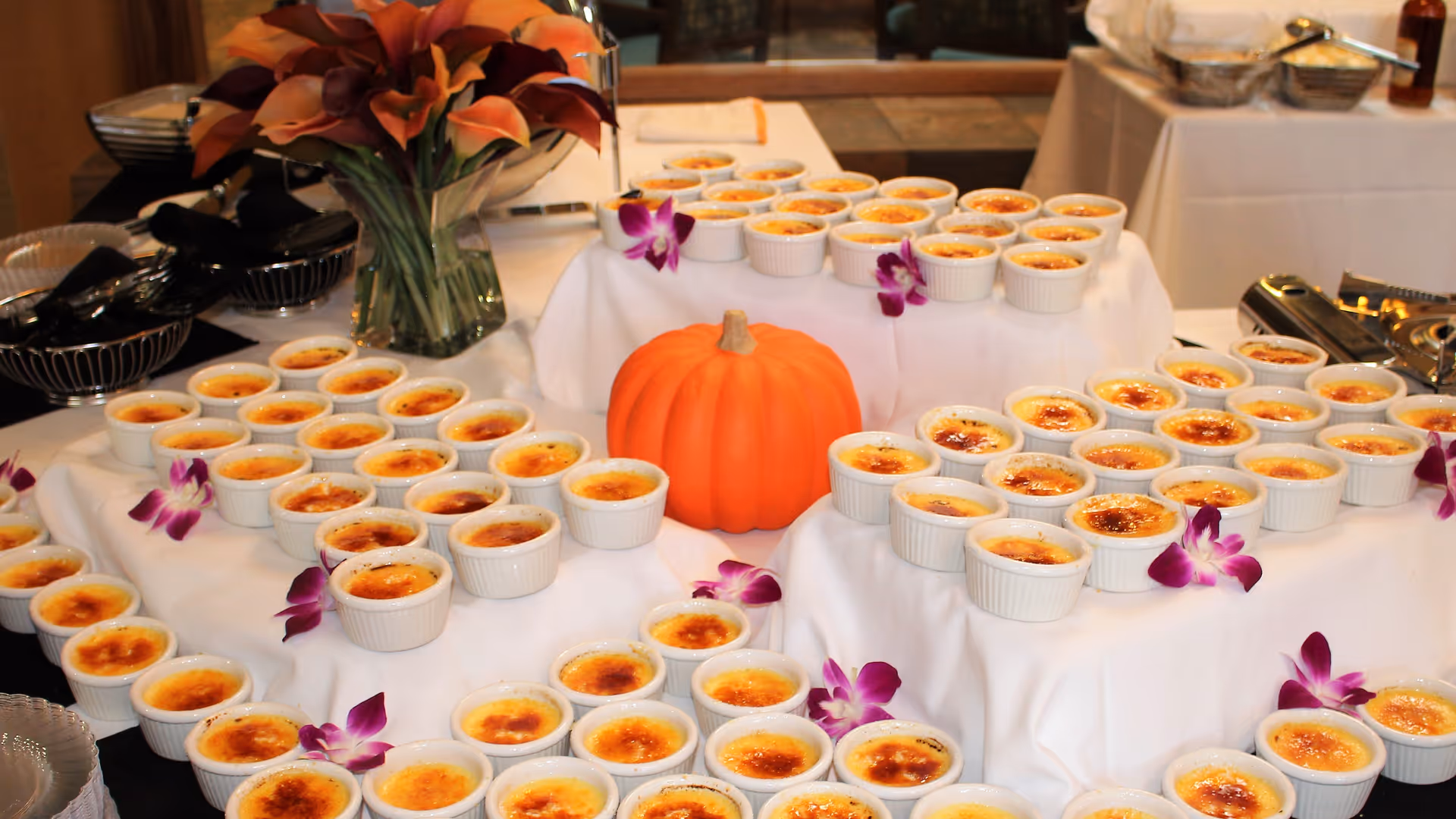 Buffet table with numerous crème brûlée ramekins arranged on white cloth around a small decorative pumpkin and purple orchid flowers.