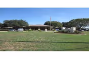 Single-story nursing facility building with a wide front lawn, flagpole, parked cars, and trees.