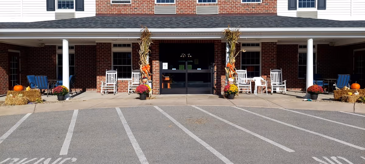 Front entrance of a brick assisted living facility with a covered porch. The porch has white columns, rocking chairs, potted flowers, hay bales, and pumpkins arranged as fall decorations. There is a paved parking area with marked parking spaces in front of the entrance.