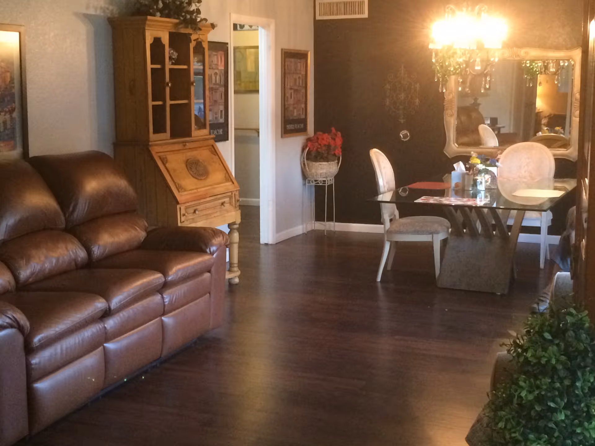 Interior view of a living and dining area in a senior living facility. The room features a brown leather sofa on the left, a wooden cabinet with glass doors, and a glass dining table with four upholstered chairs. A large mirror and a chandelier with lights are on the wall behind the dining table. The floor is dark wood, and there are decorative plants and framed pictures on the walls.