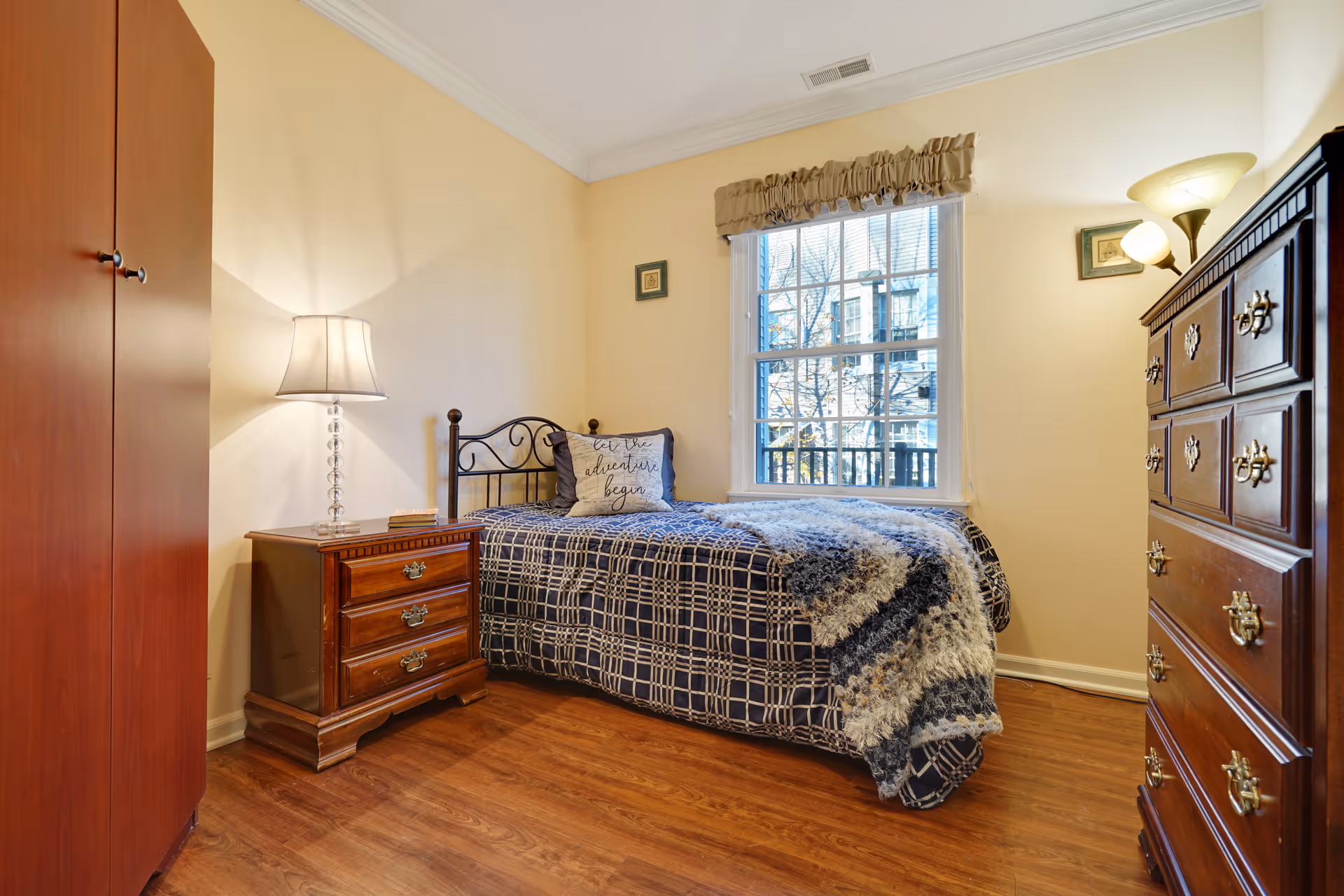 A cozy bedroom with a single bed covered in a blue and white checkered blanket and a fluffy throw. There is a decorative pillow on the bed with the words 'let the adventure begin'. The room has wooden flooring, a wooden nightstand with a lamp, a wooden dresser, and a window with beige curtains letting in natural light.