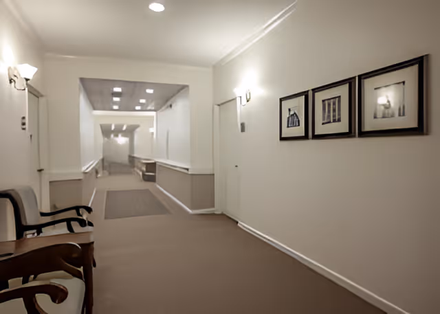 Well-lit neutral hallway in a senior living facility with seating along the left wall, framed artwork on the right, and a long corridor ahead.