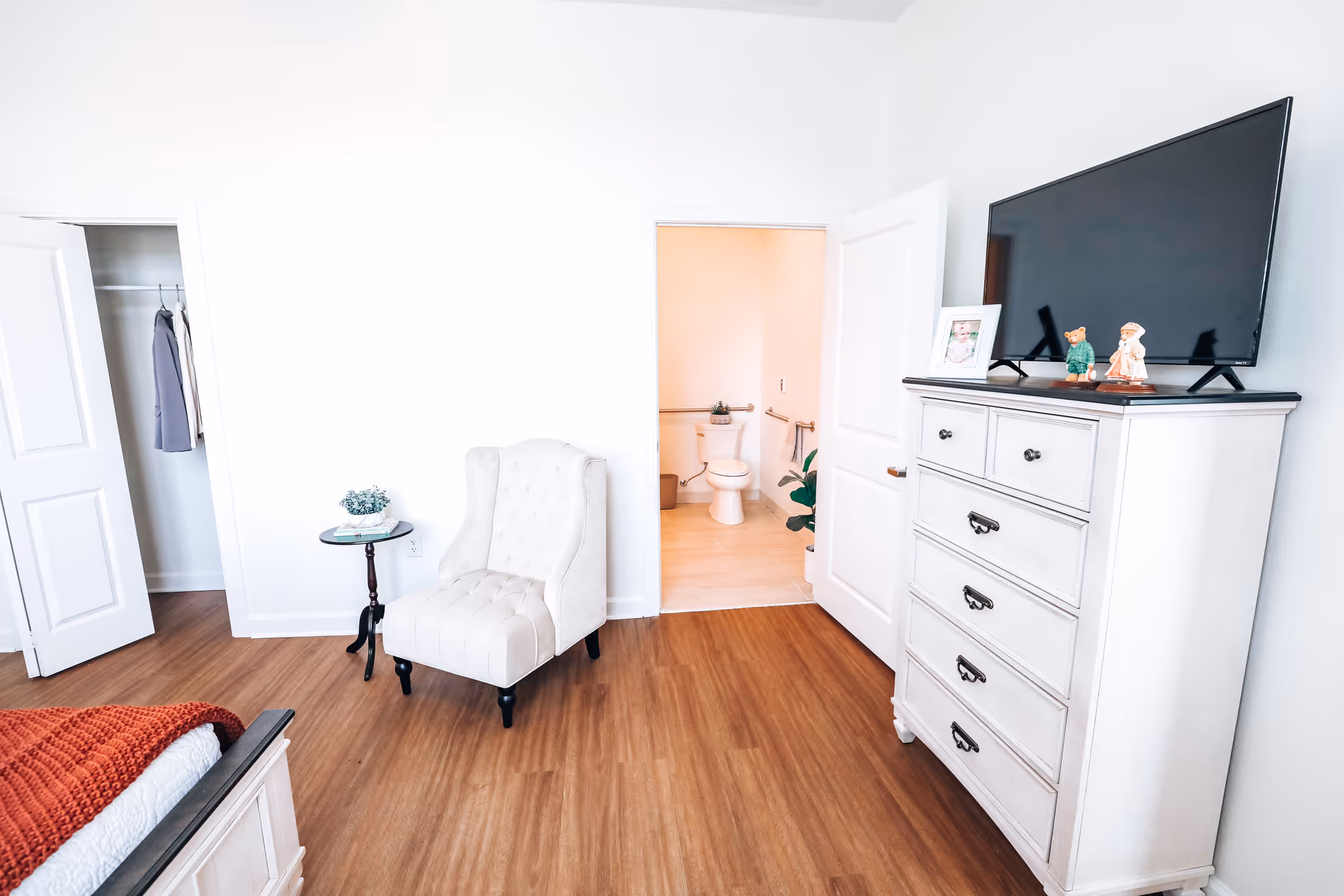 Bright bedroom with a tufted chair, white dresser topped by a TV, and an open door revealing an adjacent bathroom with a toilet.
