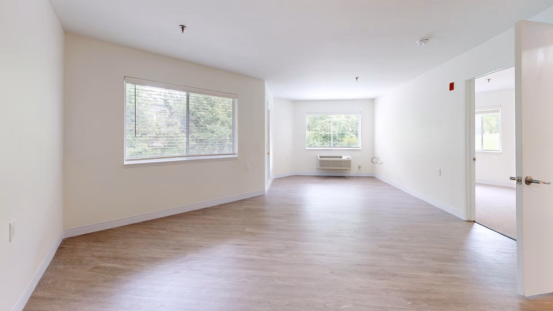 Empty room with light-colored walls and wood flooring, two large windows with blinds letting in natural light, an air conditioning unit below one window, and an open door leading to another room with carpeted flooring.