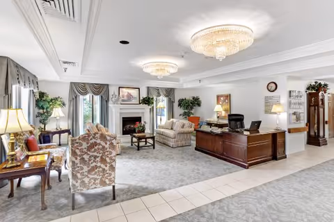 A bright and elegant senior living facility common area featuring floral upholstered armchairs, a wooden desk with a computer and lamp, a fireplace with framed artwork above it, large windows with draped curtains, potted plants, and two ornate ceiling chandeliers.