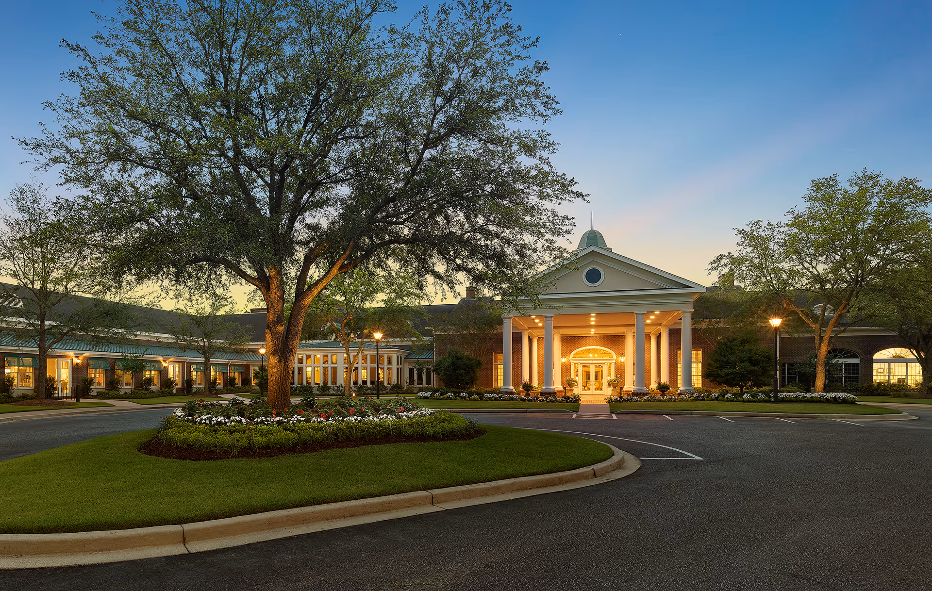 Front entrance of a well-lit retirement community building with columns, landscaped roundabout and large trees at dusk.