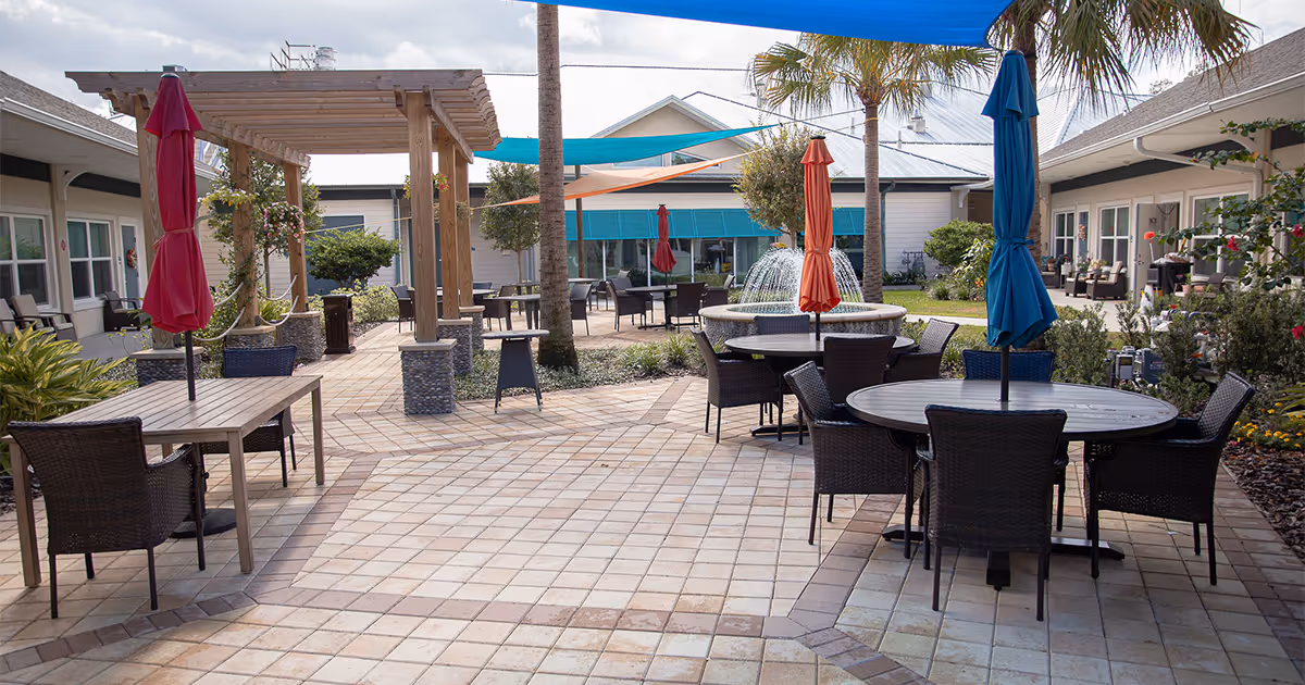 Outdoor patio area at A Banyan Residence Assisted Living Resort featuring round and rectangular tables with wicker chairs and closed umbrellas in red, orange, and blue. There is a water fountain in the center, surrounded by greenery, palm trees, and shade sails overhead. The patio is paved with light-colored tiles and is bordered by single-story buildings with windows and doors.