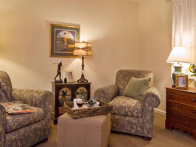 Cozy sitting area with two patterned armchairs, side tables and lamps, a small ottoman with a tray holding a glass of wine, and framed artwork on the wall.