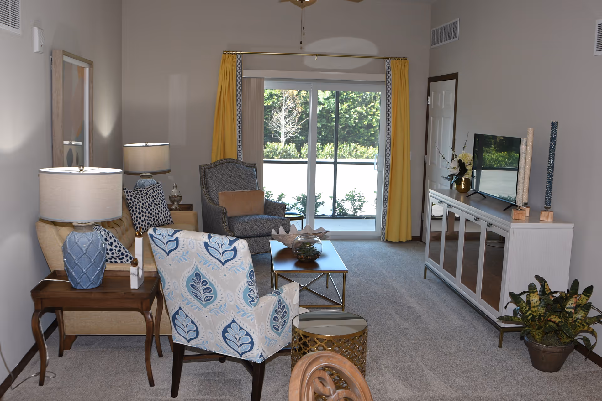 A bright living room with patterned armchairs, side tables and lamps, a coffee table, TV console, and sliding glass doors with yellow curtains.
