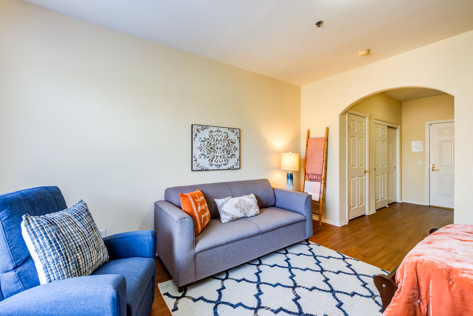 A cozy living room area with a blue armchair and a gray sofa adorned with decorative pillows. A patterned rug covers the wooden floor, and a lamp on a side table provides warm lighting. There is a decorative wall hanging above the sofa and a wooden ladder with blankets leaning against the wall near an arched doorway leading to a hallway with multiple doors.
