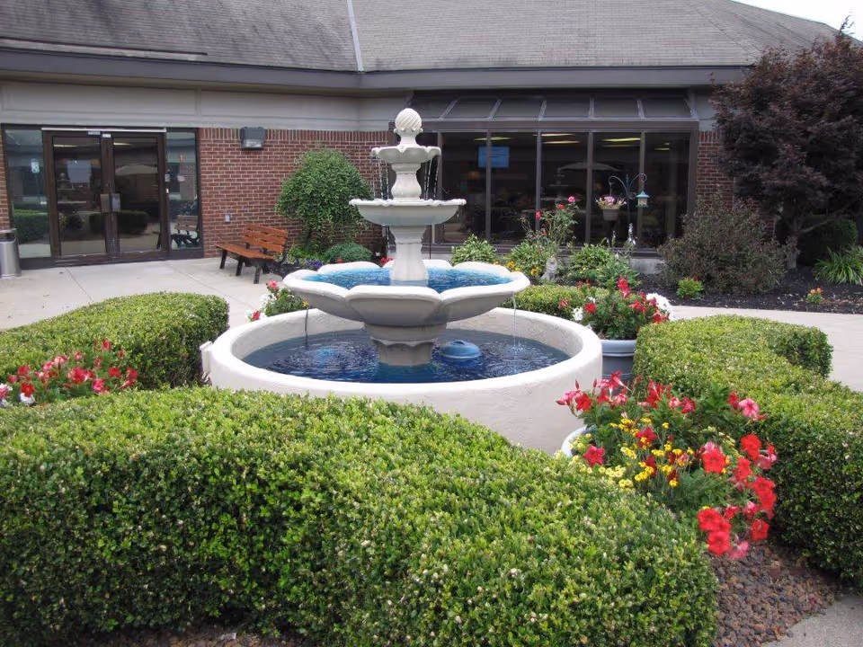 A tiered fountain surrounded by trimmed hedges and colorful flowers in front of the brick entrance to a senior living facility.