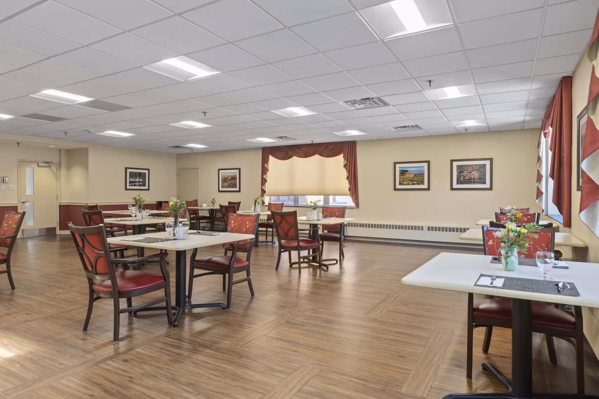 A dining room with several tables and chairs arranged neatly. Each table has a small vase with flowers, placemats, and dining utensils. The room has wood-patterned flooring, beige walls with framed pictures, and windows with red and white curtains. The ceiling has recessed lighting panels.
