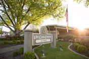 Outdoor view of the Life Care Center of Boise facility sign surrounded by landscaped greenery and trees, with the building and an American flag visible in the background during daylight.