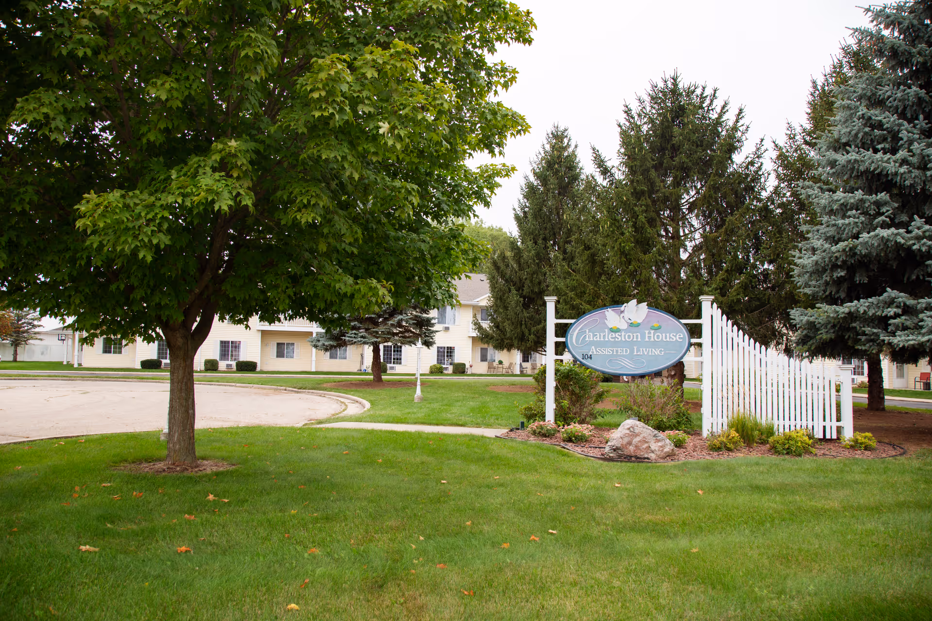 Outdoor view of Charleston House Assisted Living facility with a large green tree, manicured lawn, a white picket fence, and a sign displaying the facility's name. The building is visible in the background with multiple windows and surrounded by other trees.