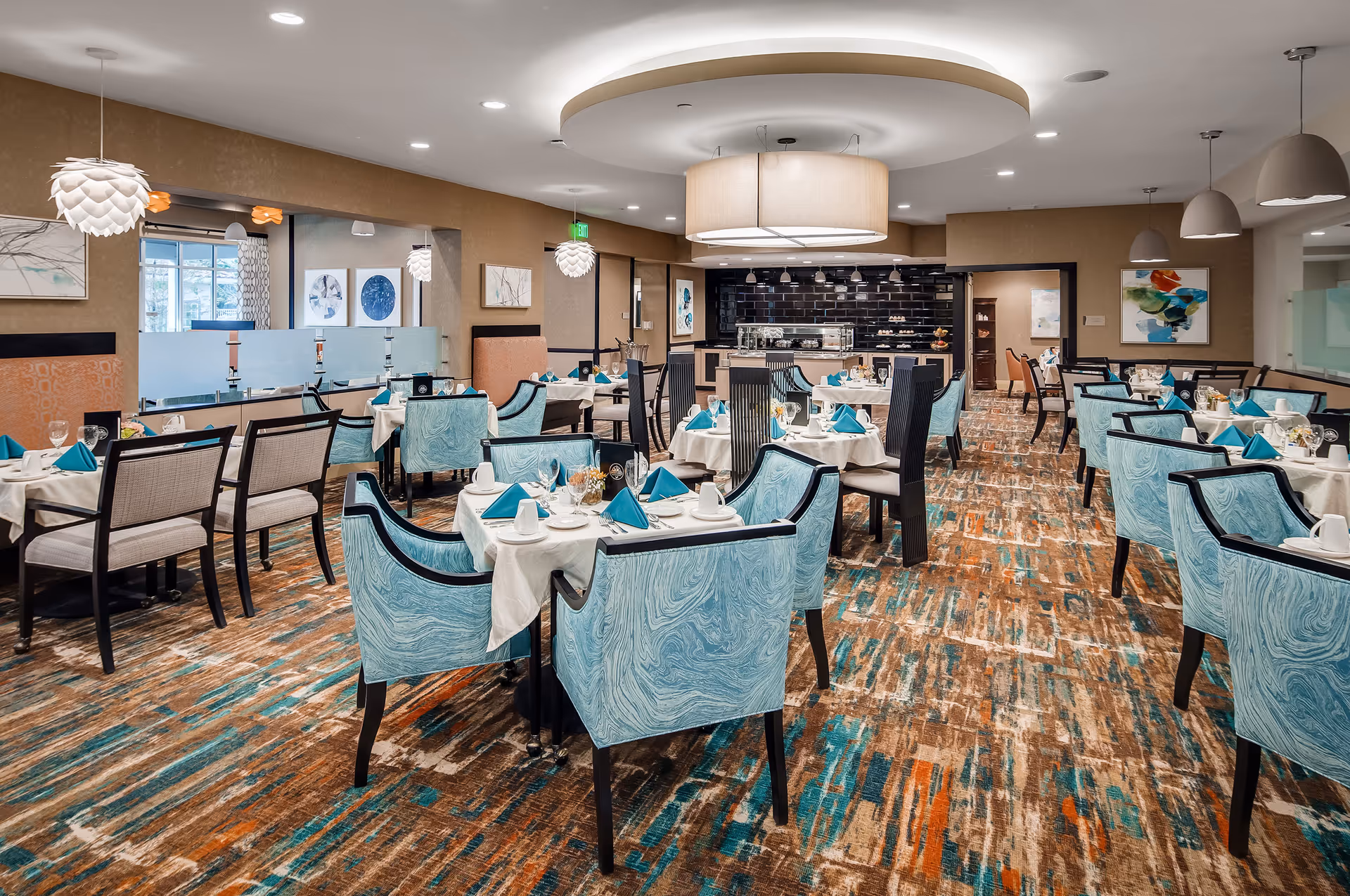 A spacious dining room with multiple tables set for a meal, featuring white tablecloths, teal napkins, and elegant tableware. The room has a patterned carpet with blue, orange, and brown tones, light blue upholstered chairs, and modern pendant lighting. Artwork decorates the walls, and a kitchen area with black tiled backsplash is visible in the background.