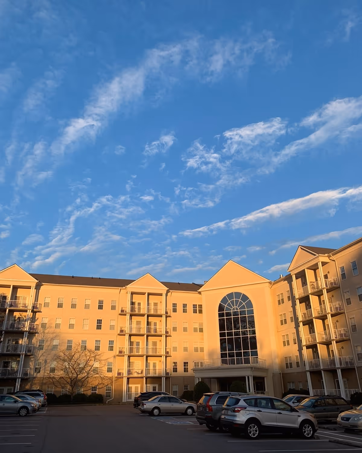 Exterior view of a multi-story senior living community building with large windows and balconies, a parking lot with several cars in front, under a blue sky with scattered clouds.