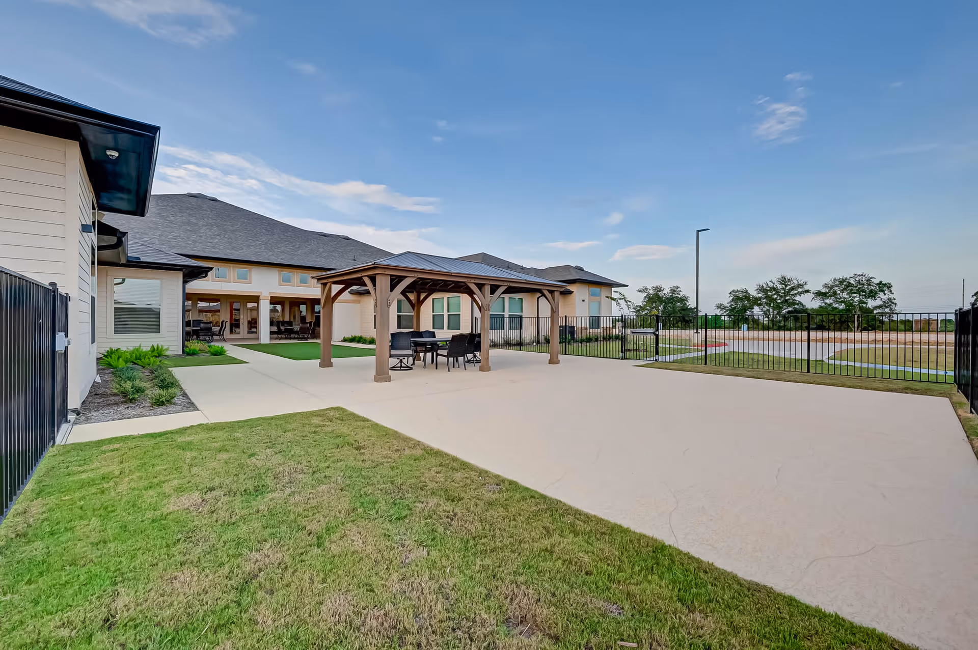 Outdoor courtyard and patio with a wooden pavilion and seating beside a fenced lawn and attached building under a blue sky.