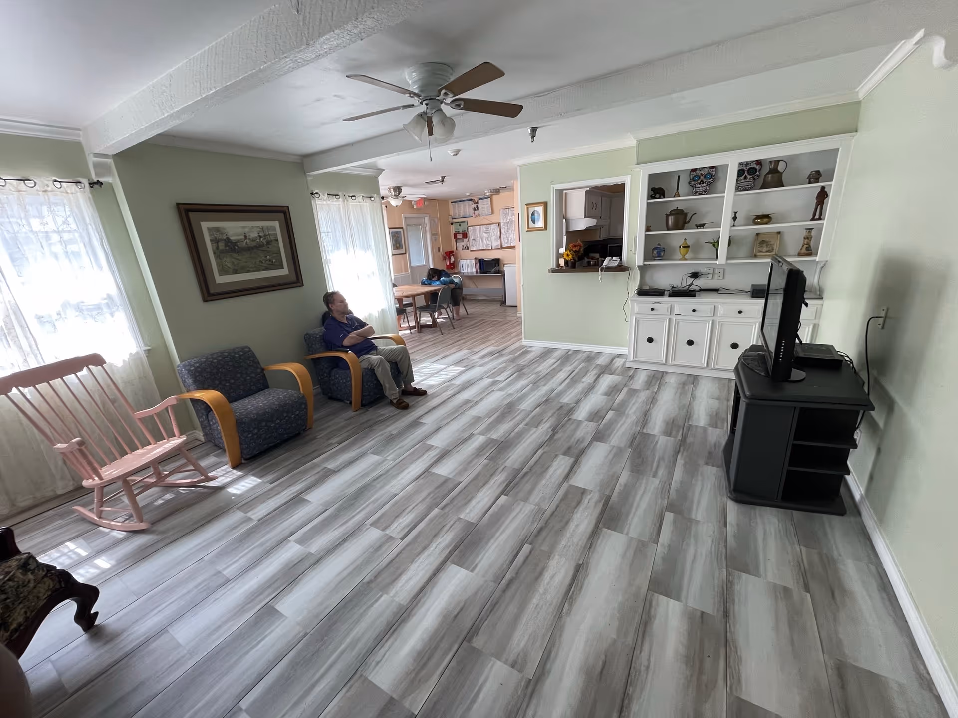 Sunlit common living room with seating including a pink rocking chair, a TV, built-in shelving, and a person seated near the windows.