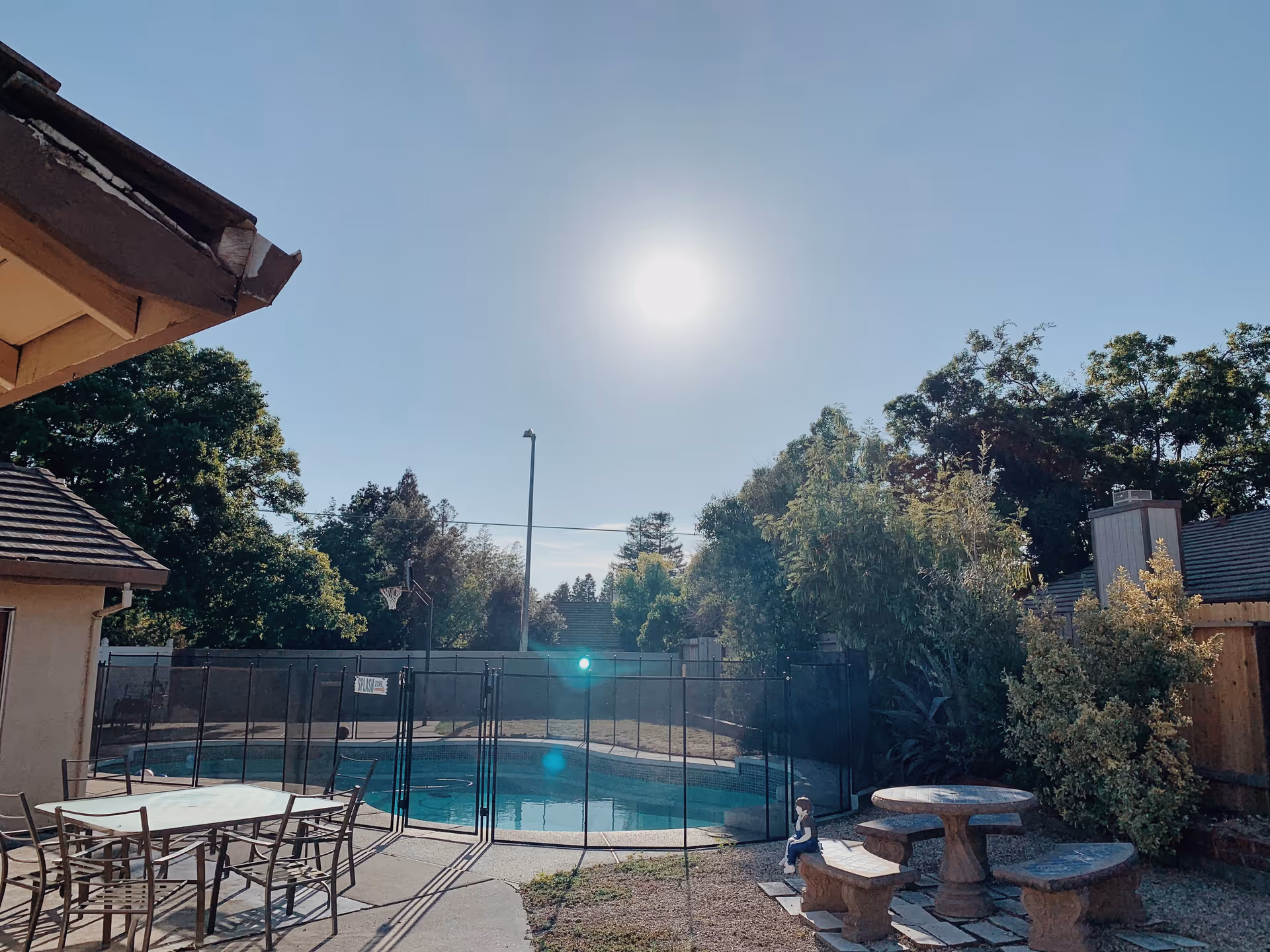 Outdoor area of a senior care home featuring a fenced swimming pool, a table with chairs, a round stone picnic table with benches, surrounded by trees and bushes under a clear sky with the sun shining.
