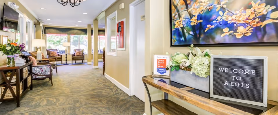 Interior view of a senior living facility hallway with beige walls and patterned carpet. On the right, a wooden console table holds a flower arrangement, a sign that reads 'WELCOME TO AEGIS', and a small framed certificate. Further down the hallway, there are chairs and tables near large windows letting in natural light. A flat-screen TV is mounted on the left wall above a small table with a flower vase.