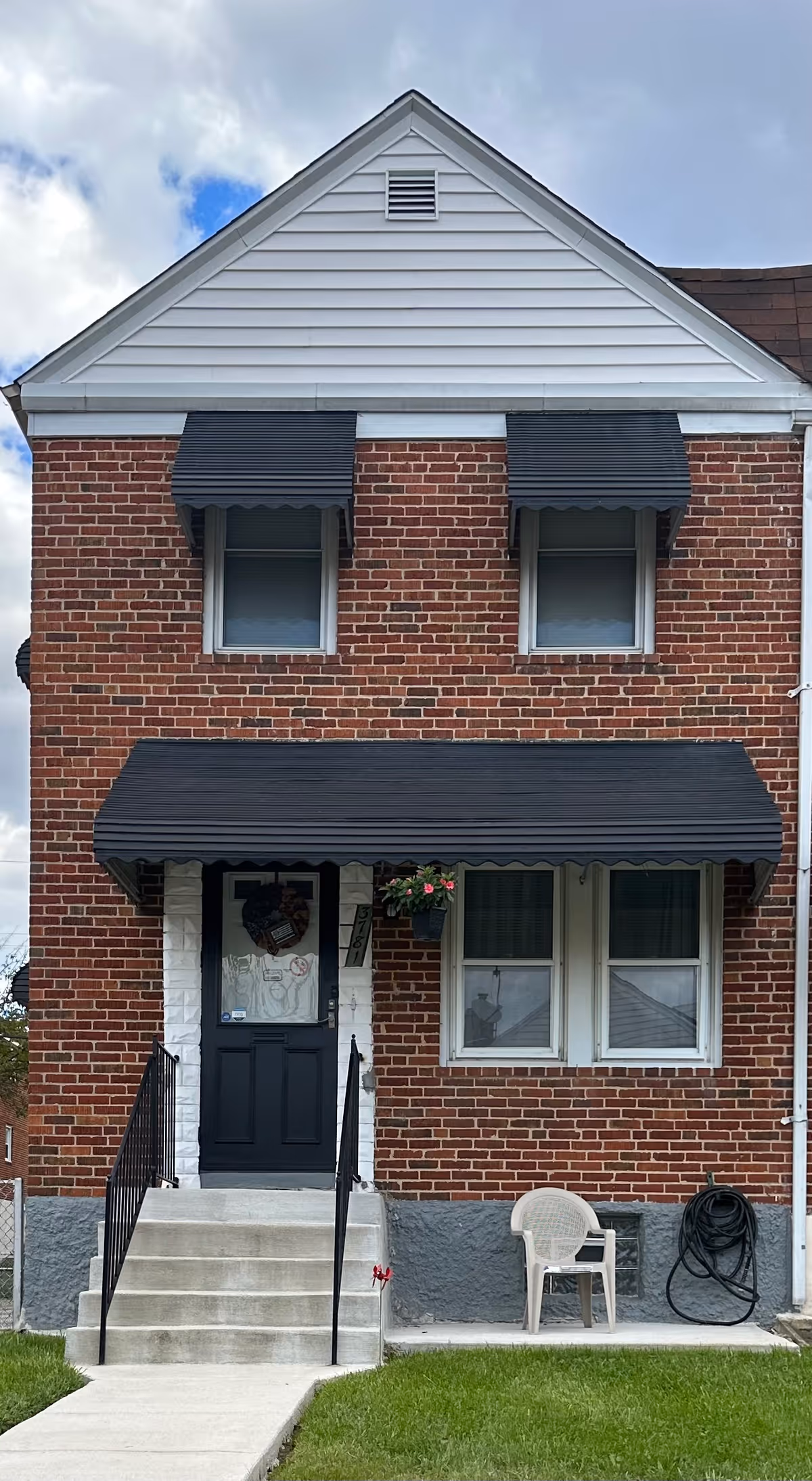 Brick two-story building with black awnings, front door, stairs, and a plastic chair outside.