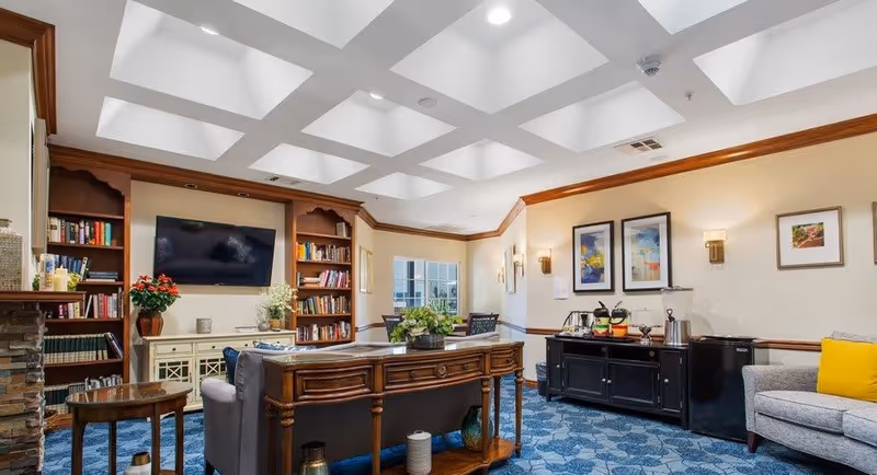 A cozy living room area with a blue patterned carpet, a gray sofa with a yellow cushion, a wooden console table behind the sofa, and built-in bookshelves flanking a wall-mounted flat screen TV. The room has a coffered ceiling with recessed lighting, framed artwork on the walls, and a sideboard with coffee and tea supplies.