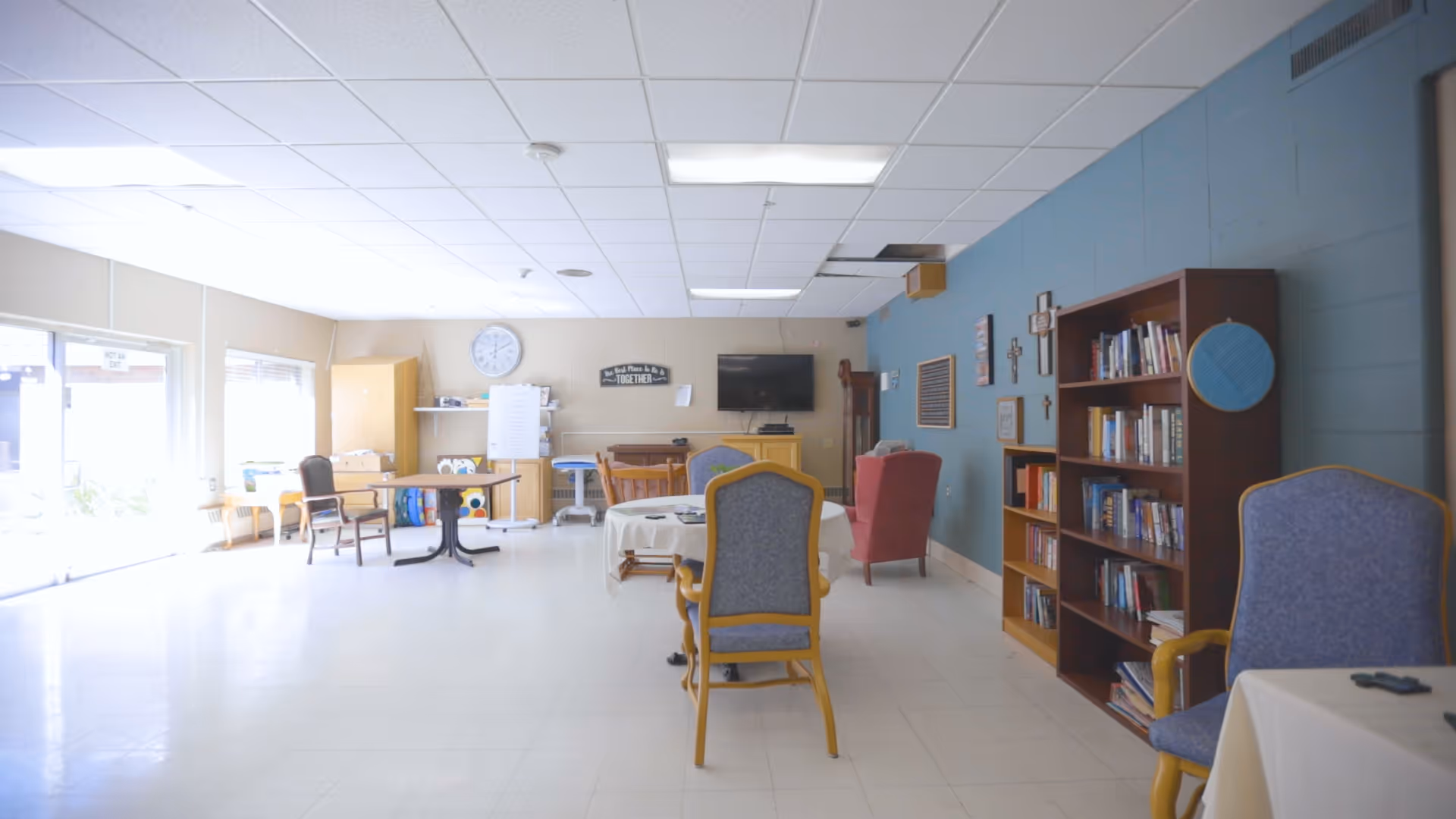 A bright common room in a senior living facility with tables and chairs arranged for seating. There is a bookshelf filled with books against a blue wall, a TV mounted on the wall, and a clock above a cabinet. The room has large windows letting in natural light and a few armchairs for comfortable seating.