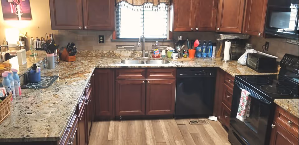 A kitchen with dark wooden cabinets, granite countertops, a double sink under a window with blinds, a black dishwasher, a black electric stove with a floral towel hanging on the handle, and various kitchen items such as utensils, water bottles, and cleaning supplies on the counters.