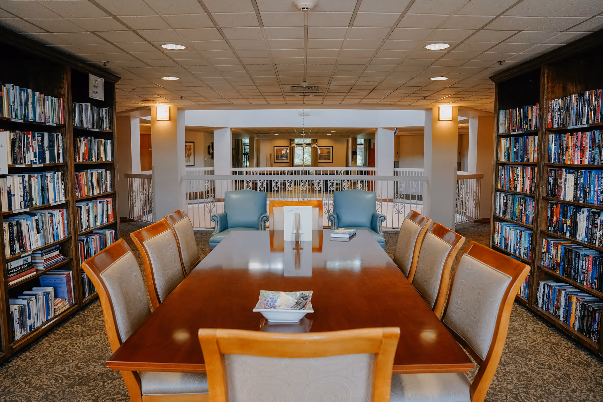 A quiet library or reading room with a long wooden table surrounded by eight cushioned chairs. On the table are a few books and a small decorative dish. The room has bookshelves filled with books on both sides and two blue armchairs at the far end near a railing overlooking another area. The ceiling has recessed lighting and the carpet has a patterned design.