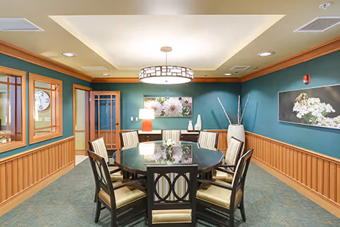 A well-lit interior room with a round glass-top table surrounded by eight cushioned chairs. The walls are painted teal with wooden wainscoting and decorated with floral artwork. A modern circular ceiling light fixture hangs above the table, and there is a lamp on a side table against the wall.