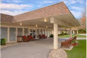 Covered entrance area of a senior living facility with a sloped roof, several red cushioned chairs arranged along the walkway, and a well-maintained lawn with trees in the background under a partly cloudy sky.