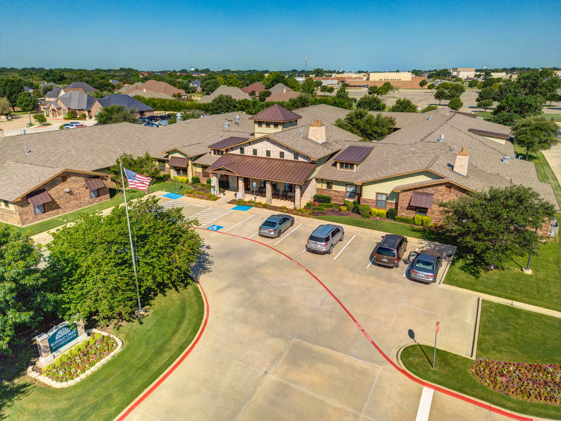 Aerial view of Buffalo Creek Assisted Living and Memory Care facility showing a single-story building with a brown roof, stone and brick exterior, a covered entrance, several parked cars, an American flag on a flagpole, and surrounding greenery and trees under a clear blue sky.
