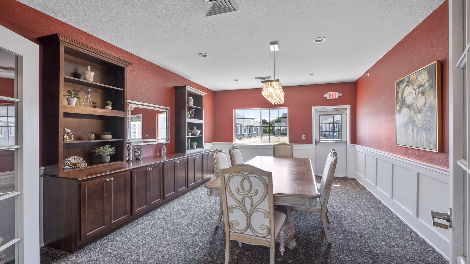 A dining room with a long wooden table surrounded by six upholstered chairs. The walls are painted red with white wainscoting, and there are two large windows and a door at the far end. A crystal chandelier hangs above the table, and built-in dark wood cabinets with shelves displaying decorative items line one wall. A large floral painting is hung on the right wall.