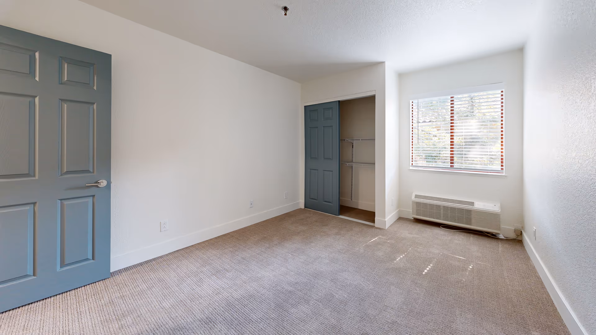 Empty bedroom with beige carpet, white walls, a window with blinds, a heating/cooling unit below the window, a closet with a sliding blue door, and a blue entry door.