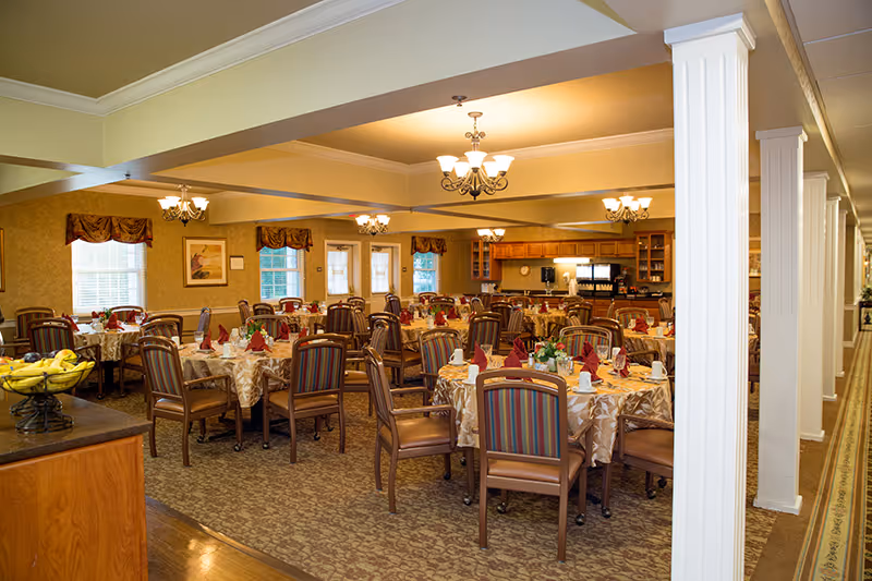 A spacious dining room in a senior living facility with multiple round tables covered with beige tablecloths, each set with napkins, cups, and silverware. The chairs have striped upholstery and wooden frames. The room is warmly lit with chandeliers and has windows with valances. A kitchen area is visible in the background.