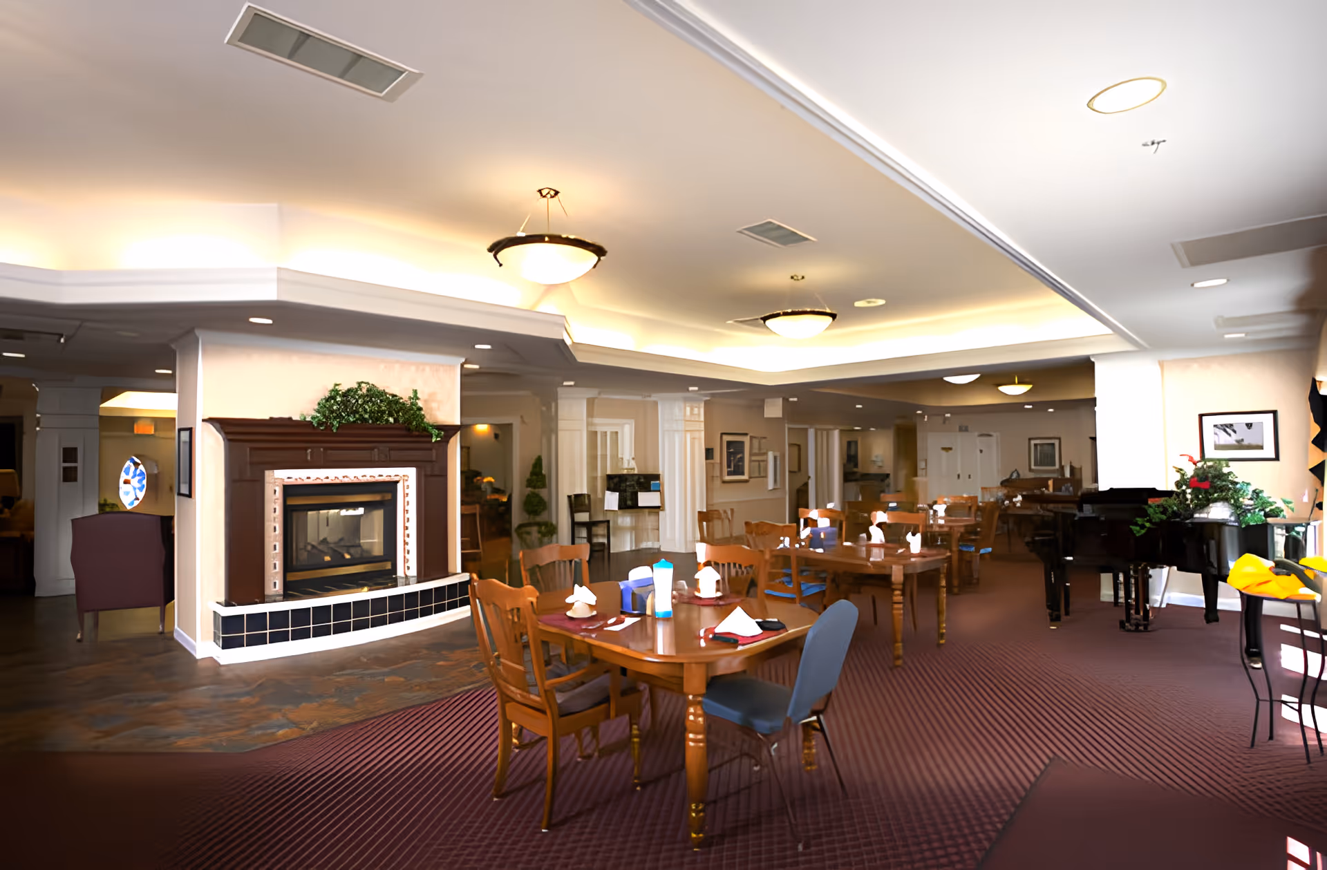 A spacious dining room in a senior living facility with multiple wooden tables and chairs arranged neatly. Each table is set with napkins and condiments. There is a fireplace with a decorative mantle and greenery above it on the left side. The room has a carpeted floor, soft ceiling lighting, and a piano near the windows on the right side. The overall ambiance is warm and inviting.