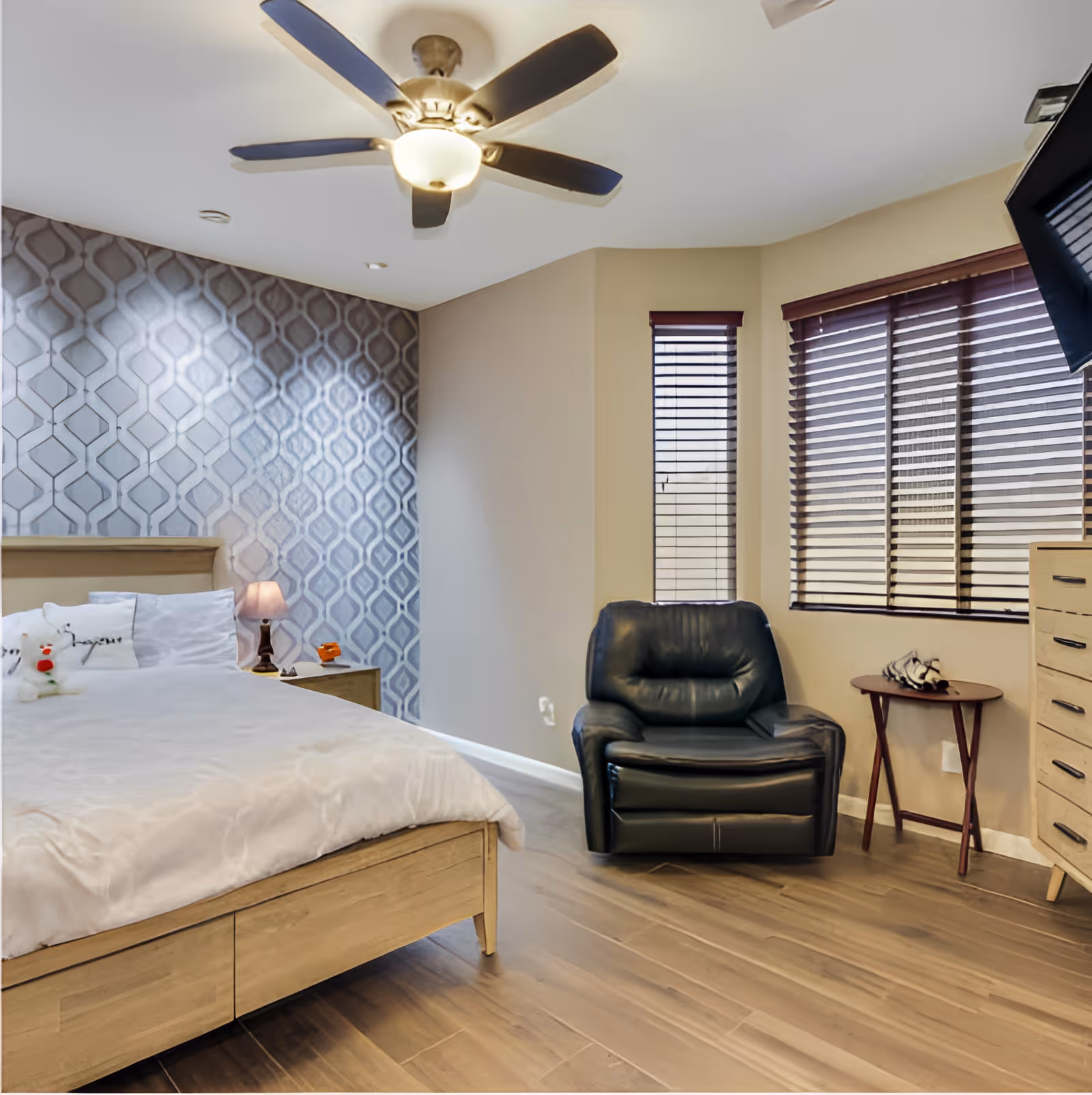 Well-lit bedroom with a bed, patterned accent wall, a leather armchair, ceiling fan, and wooden blinds.