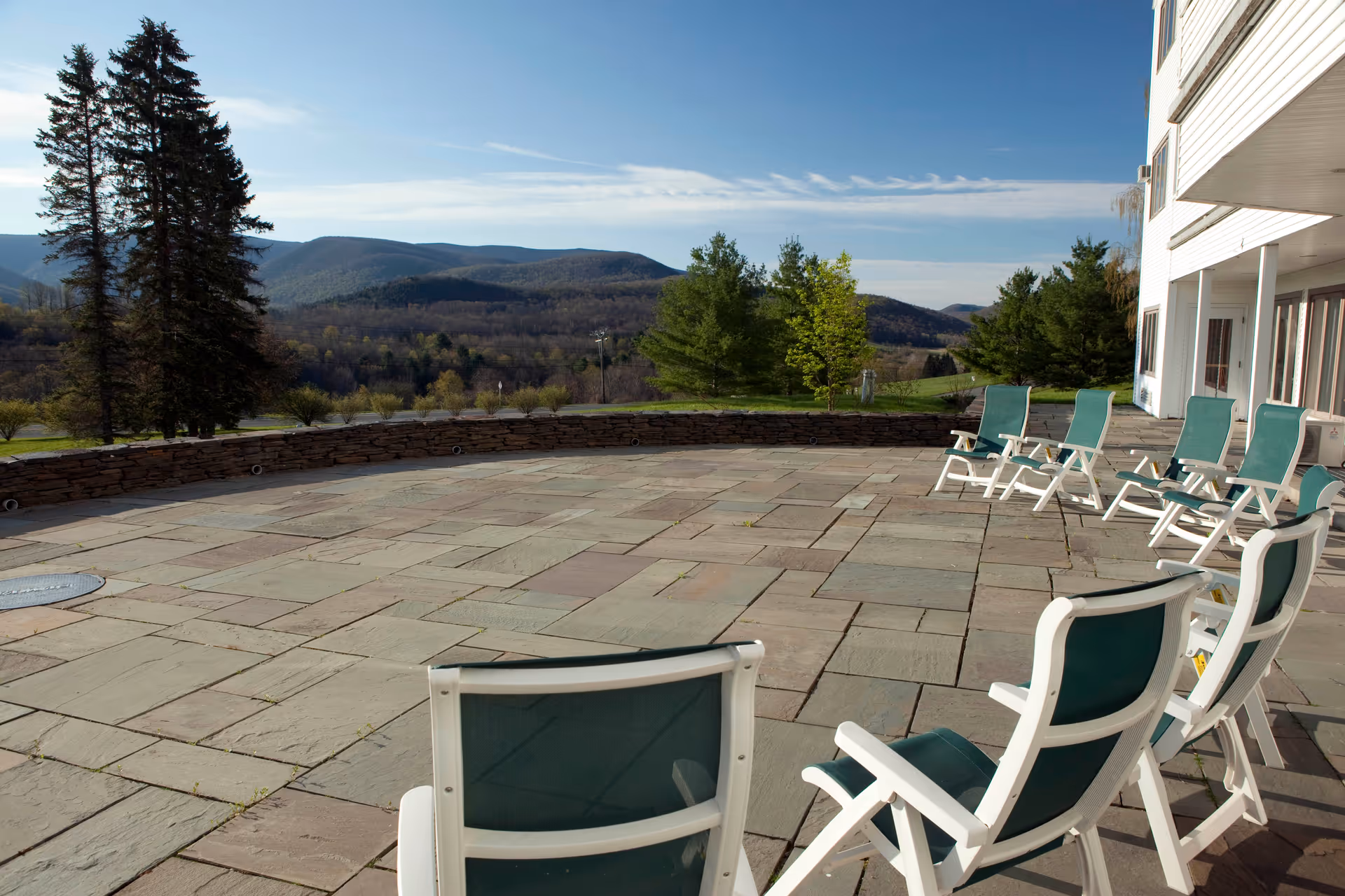 Outdoor patio area with several white-framed chairs with green fabric seats arranged around the edge, overlooking a scenic view of trees and distant mountains under a clear blue sky.