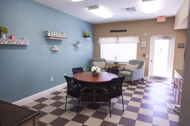 A cozy sitting area in a senior living facility with a round wooden table surrounded by four black chairs. Two patterned armchairs with cushions are placed near a window with blinds and a valance. The walls are painted teal and beige, decorated with small shelves holding plants and decorative items including a sign that says 'ANGELS'. The floor has a checkered pattern in brown and beige tiles. A door with a window and an exit sign is visible in the background.