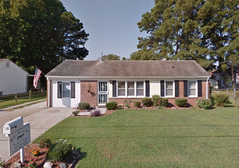 Single-story brick and siding building with a front lawn, small shrubs, and an American flag on a pole near the driveway. A sign in front reads 'Bethel Helping Hands Assisted Living'.