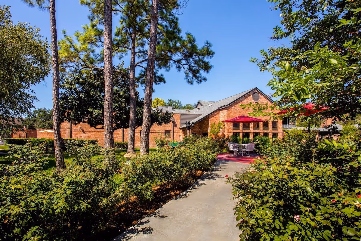 Outdoor view of a senior living facility with a paved pathway leading through lush green bushes and tall trees towards a brick building. There are red umbrellas shading outdoor tables and chairs on a patio area beside the building under a clear blue sky.