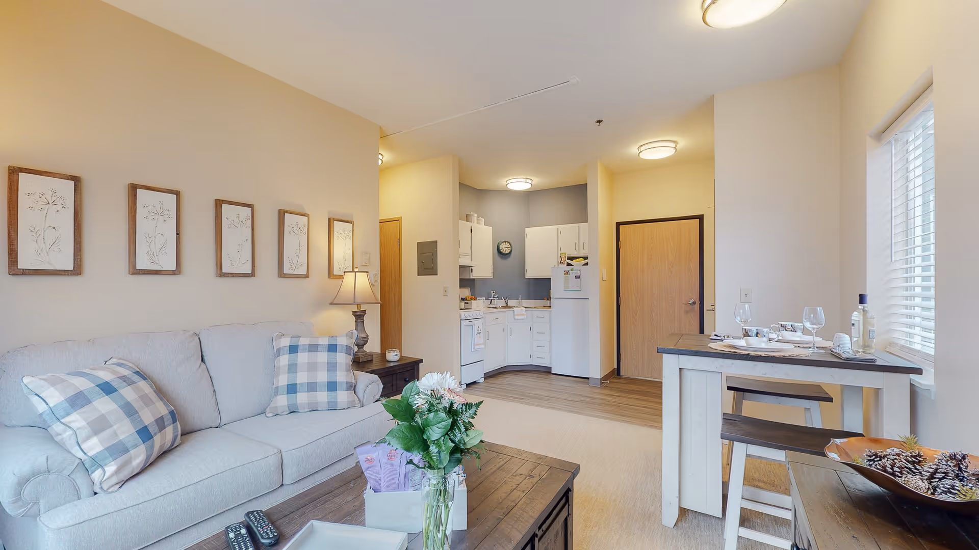 A cozy living area with a light gray sofa adorned with two blue and white checkered pillows. Above the sofa, five framed botanical prints are hung on the wall. A wooden coffee table in front of the sofa holds a vase with flowers and some snacks. To the right, there is a small dining table set for two with wine glasses and a bottle of wine near a window with blinds. In the background, a compact kitchen with white cabinets, a stove, and a refrigerator is visible. The room has warm lighting and a neutral color palette.