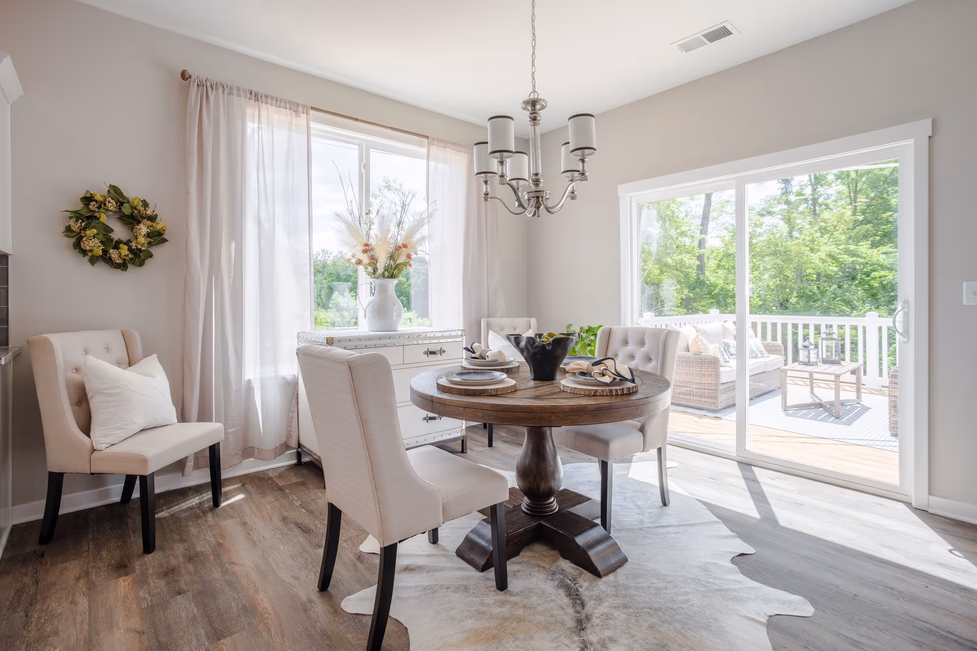 Bright dining room with a round wooden table and upholstered chairs opening to a sunlit deck through sliding glass doors.